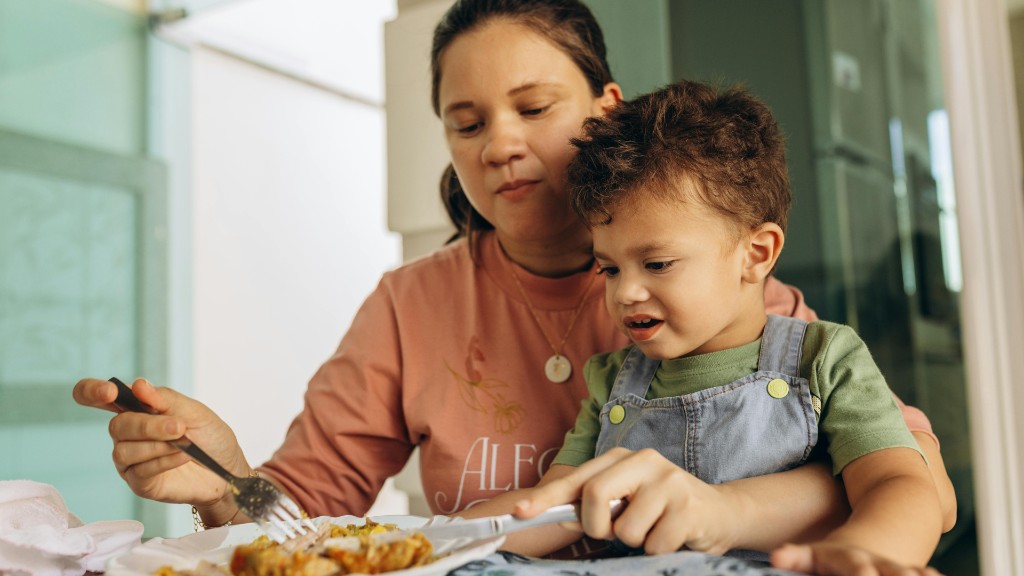 A mother sitting closely with her young son at the table, gently supporting him as he learns to cut food on his plate