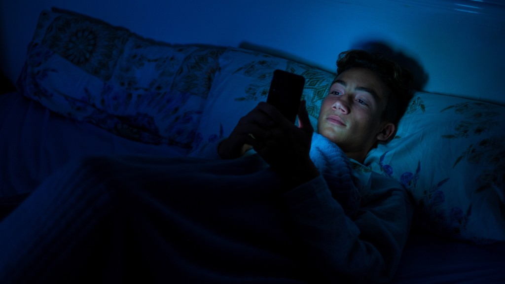A young boy lying in bed at night, his face illuminated by the glow of his smartphone as he stares at the screen