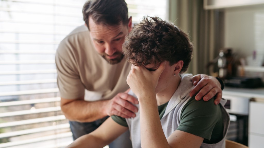 A father gently comforting his distressed teenage son, placing a hand on his shoulder as the son covers his face with his hand