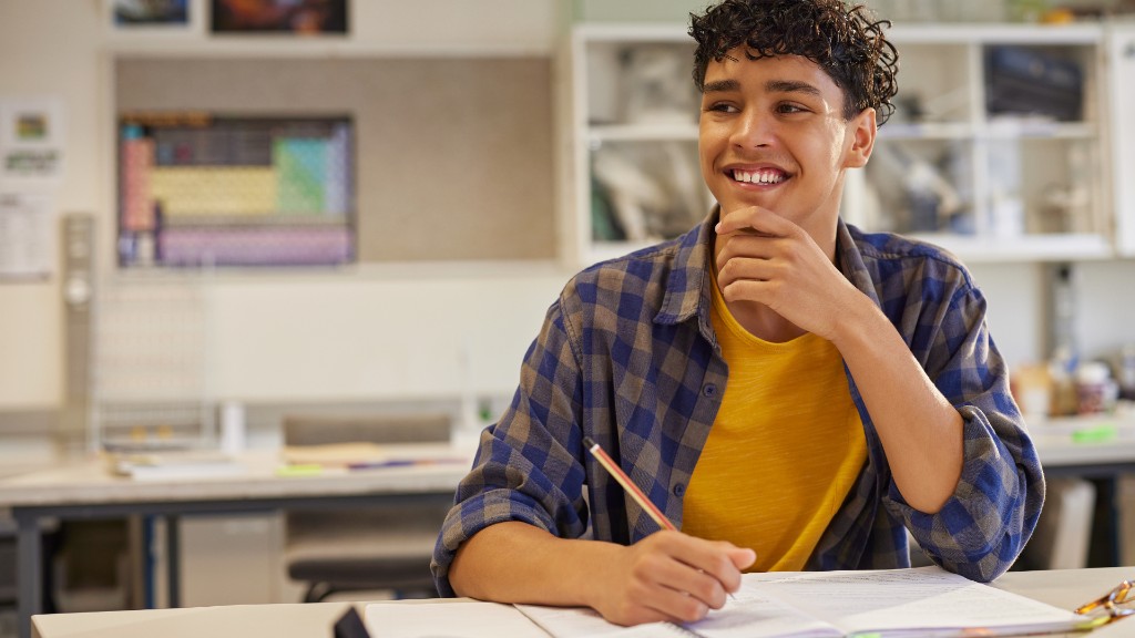 A smiling teenage boy in a classroom, resting his chin on his hand with a pencil in a thoughtful pose