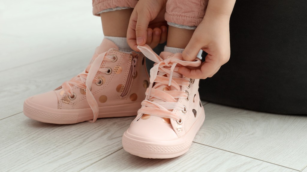 A young child sitting on an ottoman, carefully tying the pink ribbon laces on their polka-dot high-top sneakers