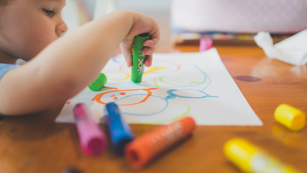 A young child drawing colorful scribbles on paper with chunky paint sticks at a wooden table