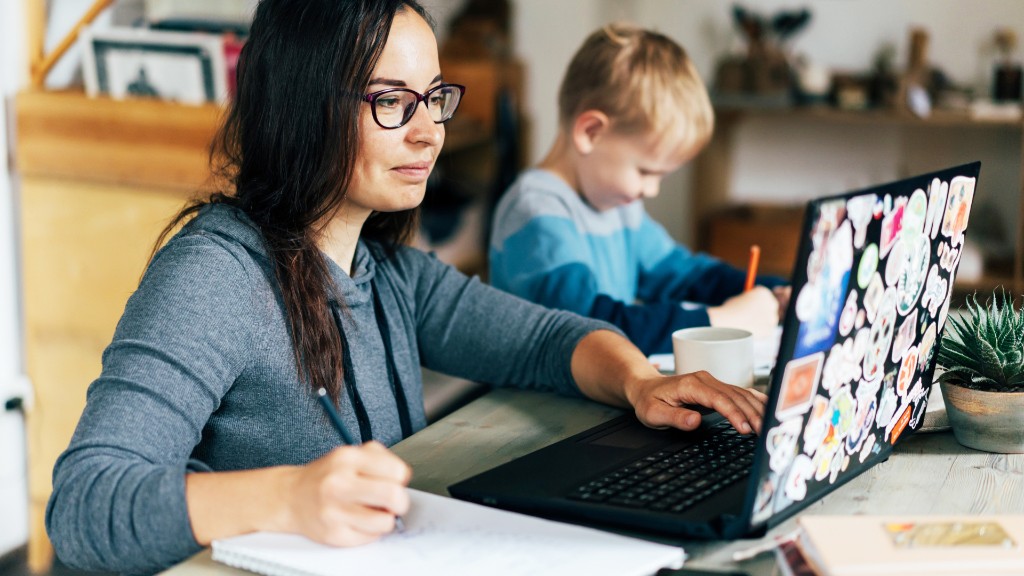 A parent working on a sticker-covered laptop at the kitchen table while a young child draws beside her