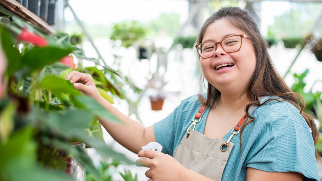 A young woman with Down syndrome smiling while caring for plants in a bright greenhouse, wearing a work apron and holding a spray bottle