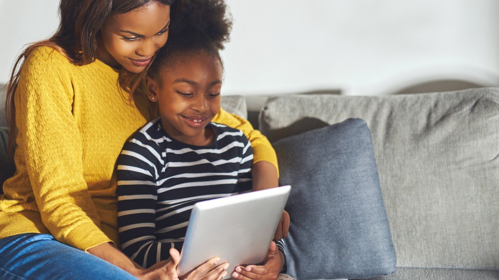 A mother and young daughter smiling together on a couch while reading on a tablet