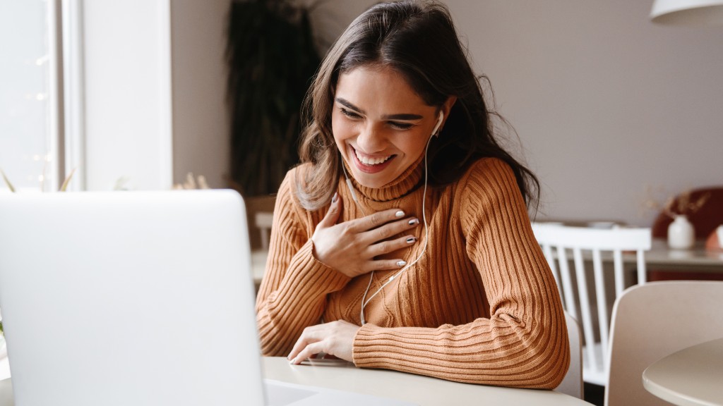 A young woman laughing warmly while on a video call on her laptop with earphones in a bright cafe