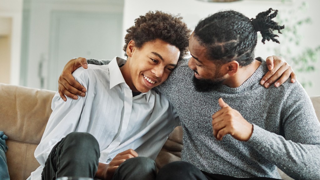 A father with his arm around his teenage son on a couch, both smiling