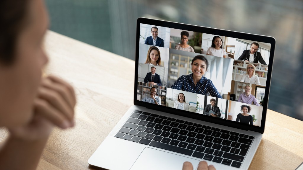 A person on a video call with a grid of participants on a laptop screen at a wooden desk