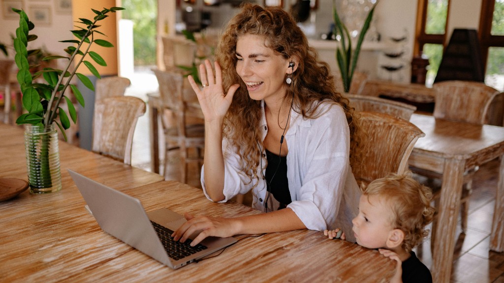 A woman smiling and waving during a video call on her laptop while a toddler peeks over the table beside her