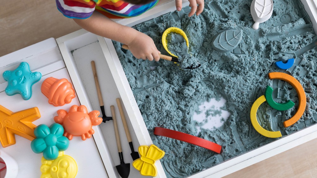 Overhead view of a child's hands using a shovel in kinetic sand with colorful molds and tools for fine motor play