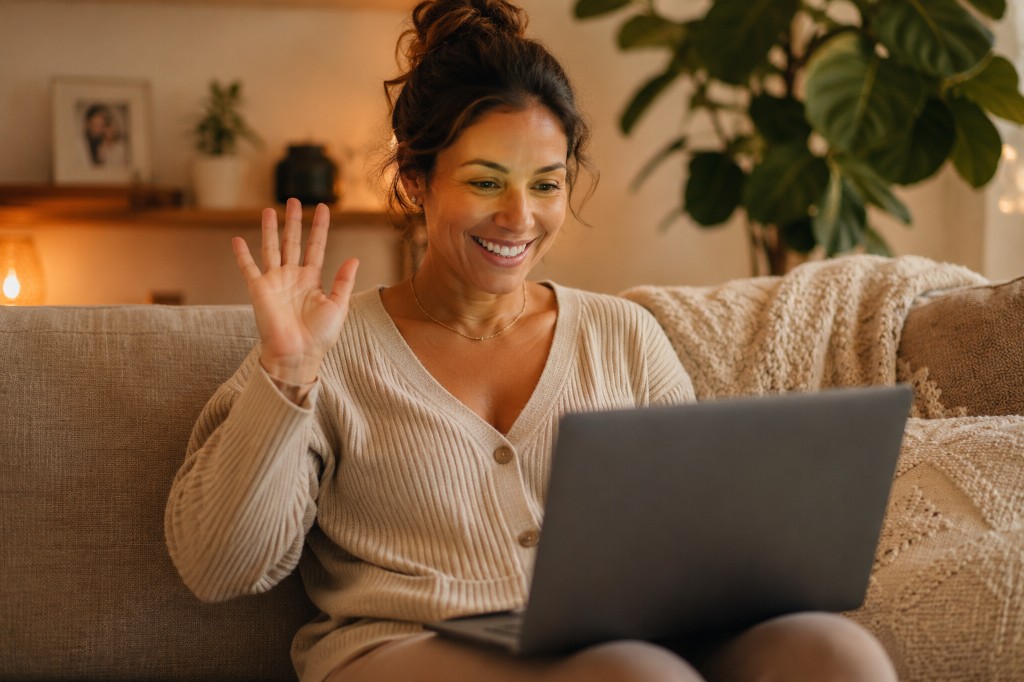 A woman smiling and waving during a video call on her laptop while sitting on a living room sofa