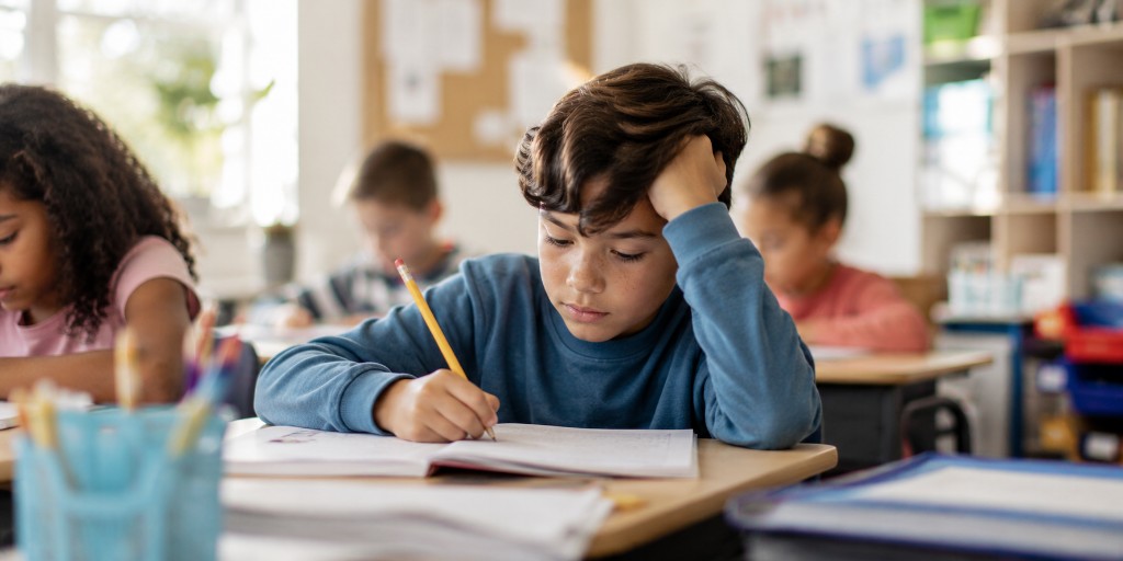 A boy leaning on his hand while writing in a notebook at a classroom desk with a pencil