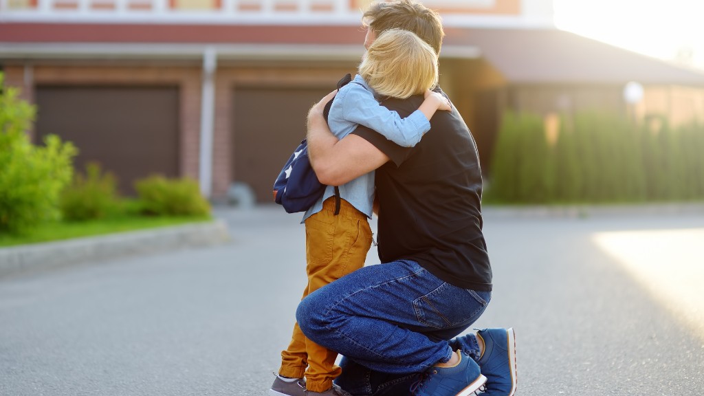 A father kneeling and hugging his young son with a backpack outside a home at golden hour