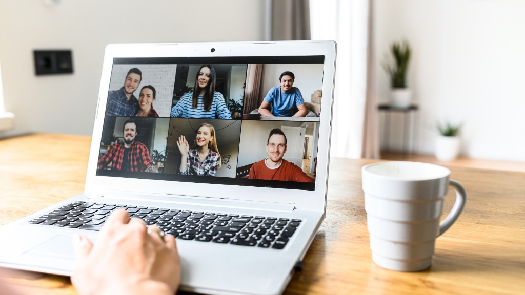 A person on a video call with six participants on a laptop screen at a wooden table