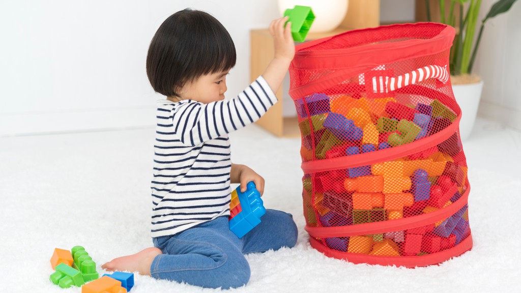 A young child carefully placing colorful building blocks into a red mesh toy basket on a white rug