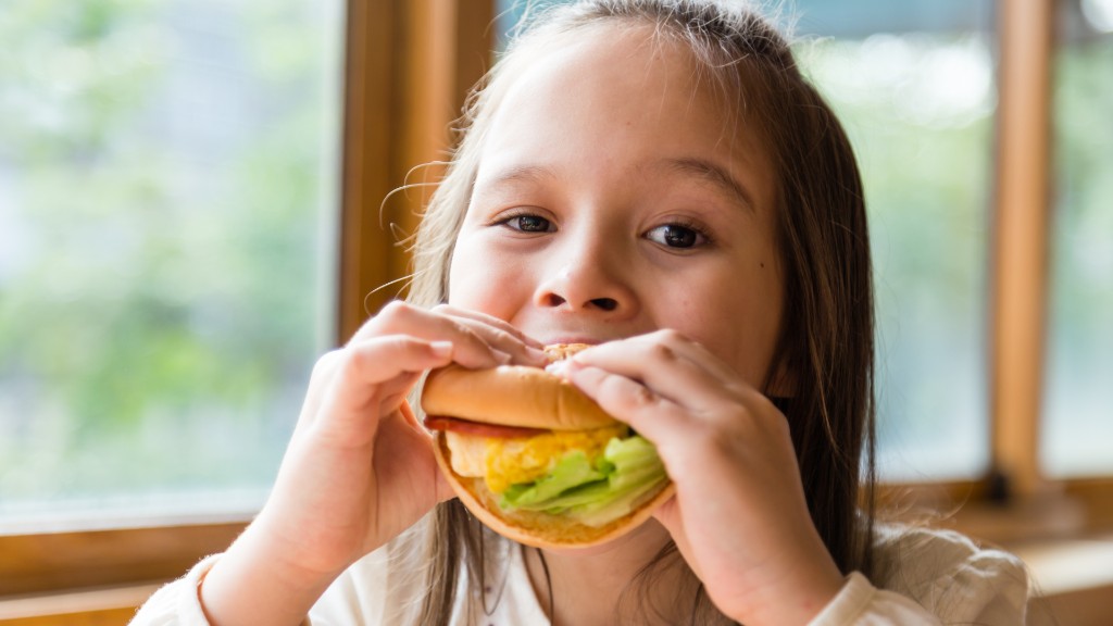A young girl happily taking a bite of a hamburger with lettuce at a bright table