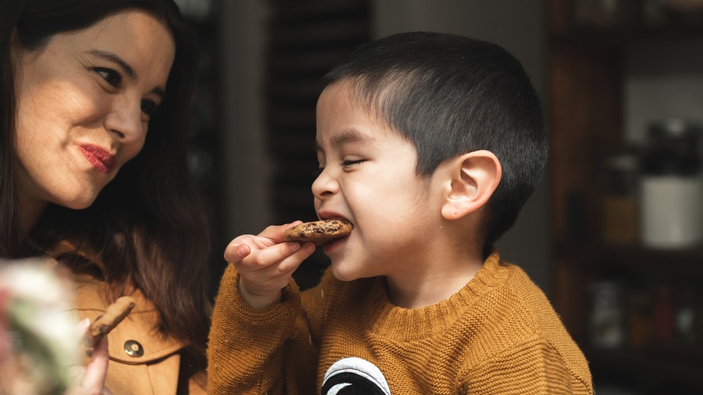 A mother smiling as her young son takes a bite of a cookie at the table
