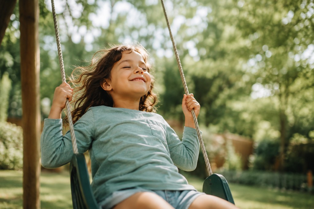 A young girl with her eyes closed and a peaceful smile while sitting on a swing in a sunlit backyard