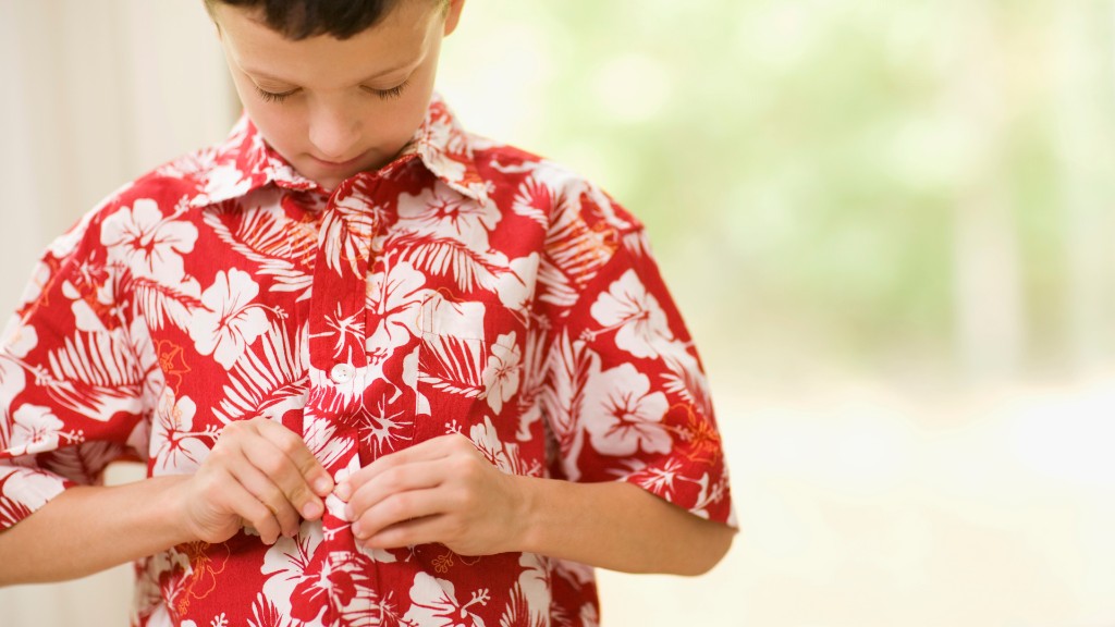 A young boy concentrating as he buttons a red tropical-print shirt, focusing on the fine motor task at his chest