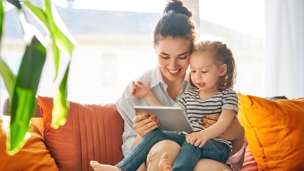A mother and young daughter sitting together on a couch, smiling while looking at a tablet