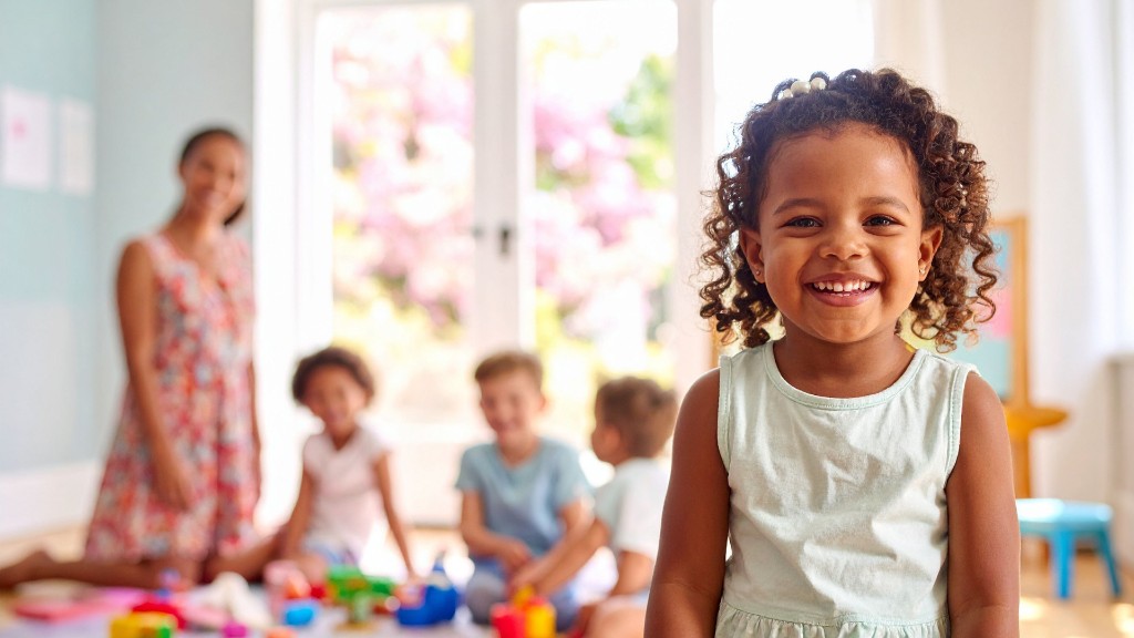 A smiling young girl in a bright classroom with a teacher and other children playing softly blurred in the background
