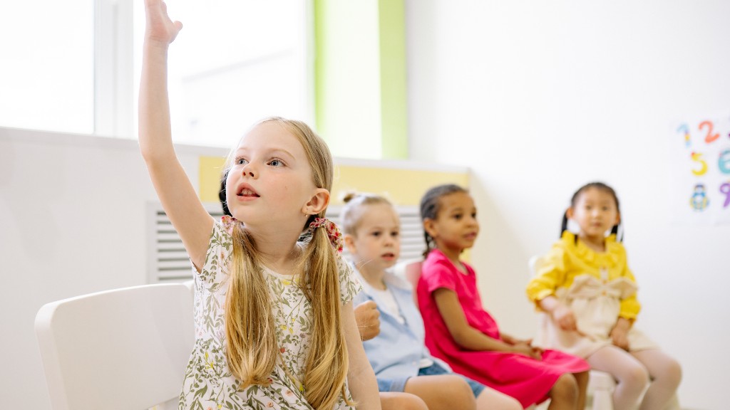A young girl raising her hand eagerly in a bright classroom with other children seated behind her