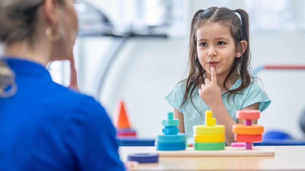 A young girl practicing a speech sound with her finger on her lip while working with a therapist across a table with colorful toys