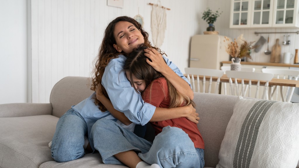 A mother embracing her daughter on a sofa as the girl buries her face in her shoulder