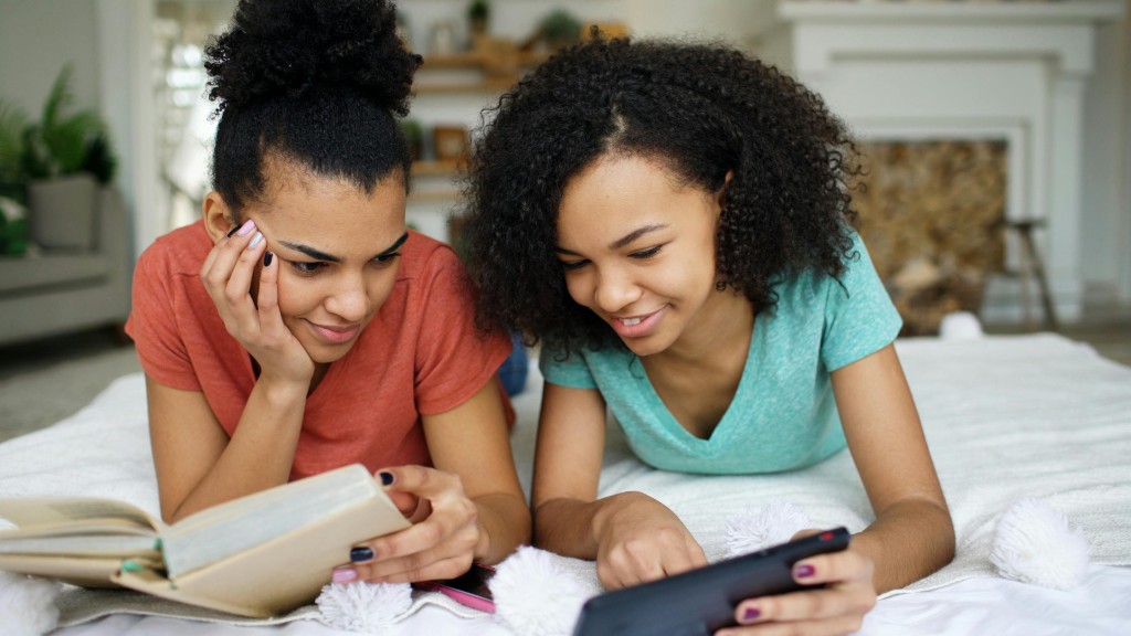 Two young girls lying on a bed together, one reading a book and the other looking at a phone