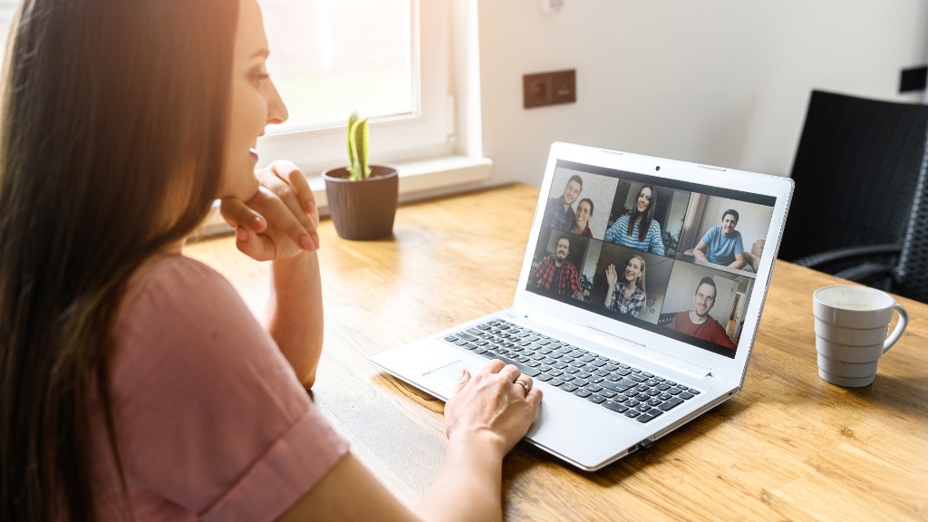 A woman on a video call with a grid of participants on her laptop screen while working at a home desk